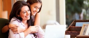 Image is of a woman smiling and working at her computer at home. Her daughter is smiling and hugging her from behind.