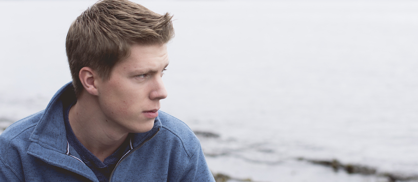 A young adult male wearing a grey-blue sweater and looking over the grey ocean.