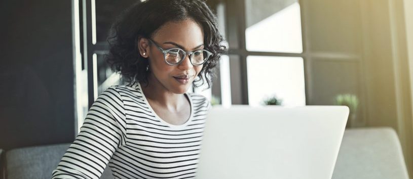 Focused young African woman wearing glasses working online with a laptop while sitting alone at a table